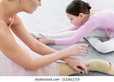 Girl Stretching On Floor In Ballet School Near Blurred Teacher