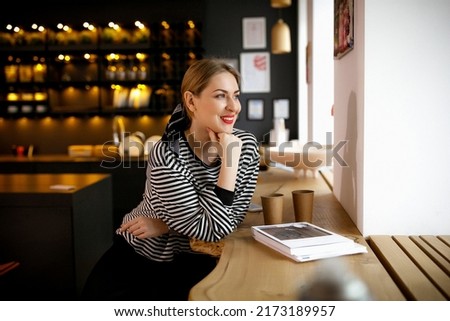 A girl in a striped sweater sits and smiles out the window in a cafe. paper cups with coffee. tea. magazine. Expecting friends. Morning mood. natural light