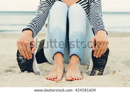 Girl in striped t-shirt and jeans sits barefoot on the beach next to the shoes