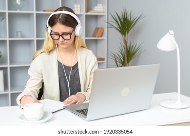 Girl student in headphones listens to the teacher's lessons online using a laptop. A young woman in glasses sitting at home writes a lecture in a notebook. Remote education.