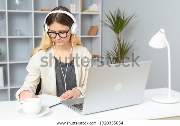Girl student in\
headphones listens to the teacher\'s lessons online using a laptop.\
A young woman in glasses sitting at home writes a lecture in a\
notebook. Remote\
education.
