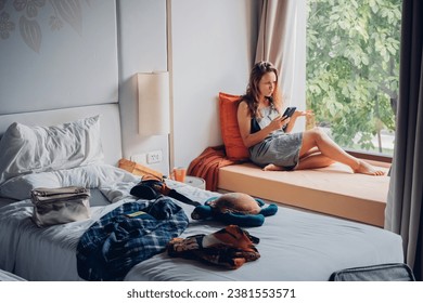 Girl student sits by the window and talks on phone in a room with a mess. Woman tourist sitting near window and talking on mobile phone in a room with mess in hotel.