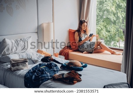 Girl student sits by the window and talks on phone in a room with a mess. Woman tourist sitting near window and talking on mobile phone in a room with mess in hotel.