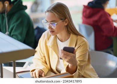 Girl student using mobile phone looking at laptop sitting at desk in university college campus classroom. Young blonde woman holding cellphone elearning on computer tech in university or coworking.