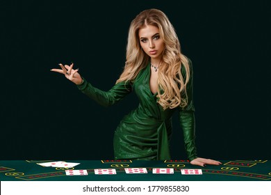 Girl in stylish dress is showing something, leaning on playing table with cards on it, posing on green background. Poker, casino. Close-up, copy space