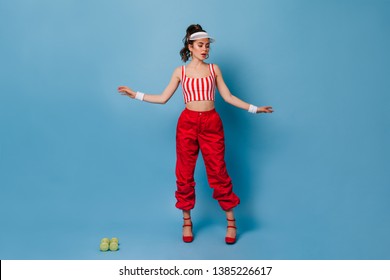 Girl In Stylish Outfit In Sporty Style Of 80s Posing On Blue Background. Portrait Of Lady In White Cap With High Tail