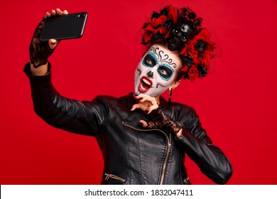 Girl with sugar skull makeup with a wreath of flowers on her head and skull, wearth lace gloves and leather jacket,making selfi isolated on red background. concept of Halloween or La Calavera Catrina.