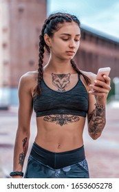 Girl in summer in city, in hand holds phone, sportswear leggings top. Pigtail hair. Tanned tattoo figure on skin. Smartphone chat online online application. Rest after workout fitness.