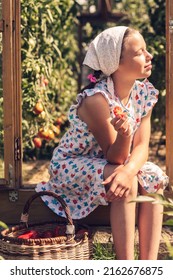A girl in a summer dress and a kerchief is eating a tomato on the threshold of a greenhouse and enjoying the summer sun.Summer and harvest concept.