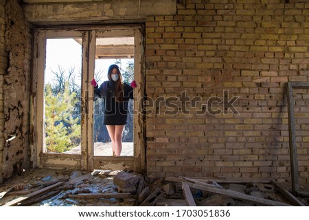 A girl in a sweatshirt stands near the door to an abandoned building in a blue medical mask and pink gloves. Coronavirus. COVID-19