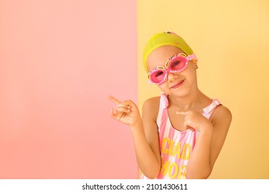 Girl swimmer in googles and swimming cap pointing with fingers aside at copy space on an pink studio background. Fashionable children's swimming goggles in the shape of a crab