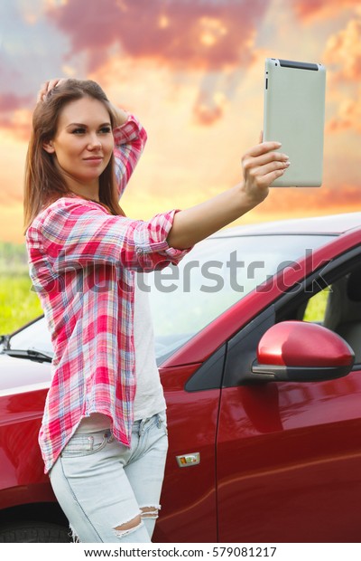 Girl with a tablet in\
hands makes selfie next to the car in the forest. Car travel,\
hitchhiking concept