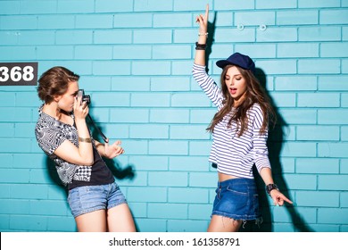 a girl takes picture of her friend in front of light blue brick wall