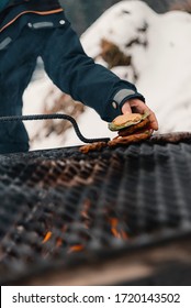 A girl taking a hamburger away from the barbeque wearing a blue ski jacket with snow in the background