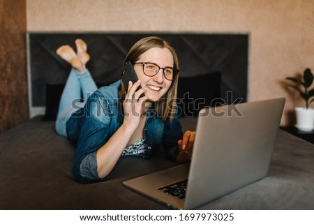 Girl talking on the phone in home. Woman working, learns and using laptop computer in the bedroom. Freelancer. Writing, typing. Communication and technology concept.