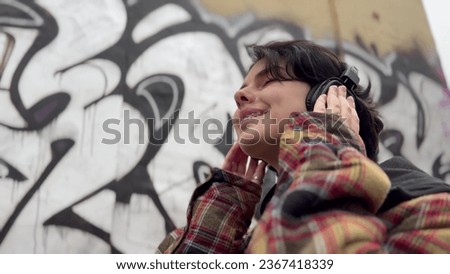 girl teenager in headphones on the background of graffiti smiles. music art freedom youth concept. brunette teenager girl walking past a wall with graffiti listening music with headphones lifestyle