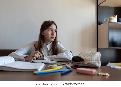 Girl thinking about school problem at table and writing solution in notebook.