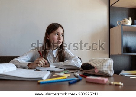 Girl thinking about school problem at table and writing solution in notebook.