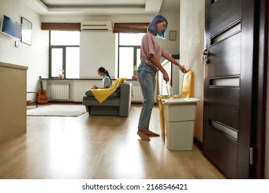 Girl Throwing Empty Plastic Bottle In Trash Can While Her Girlfriend Typing Laptop Computer On Sofa At Home. Young Caucasian Lesbain Couple. Domestic Lifestyle. Living Room In Modern Apartment