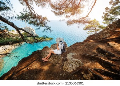 Girl tourist sits in a pine forest on the rocks on a background of beautiful seascape. View from the back.