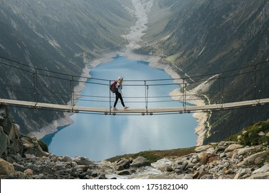 Girl Traveler With A Backpack In The Mountains. A Young Girl Stands On A Suspension Bridge On A Background Of Mountains And Glaciers. Travel And Active Life Concept. Adventure Trip To Europe.
