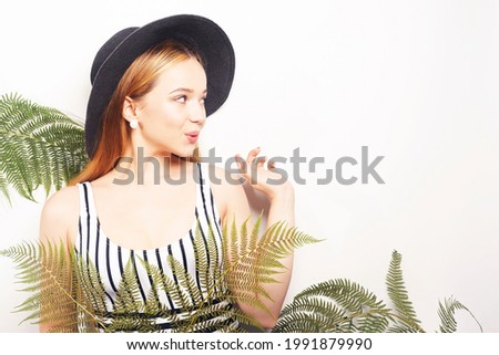 Girl in tropical leaves in a swimsuit in a big black hat smiles. Space for text, empty space point hand. Model summer photo. Big earrings