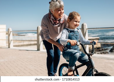 Girl trying to ride a bicycle with the help of her grandmother at seaside promenade. Senior woman holding the bicycle while her granddaughter learns to ride it.