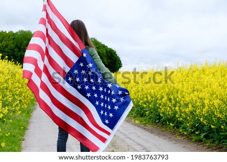 Girl turned back holding a high American flag on a field road among yellow flowers. High quality photo