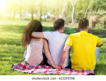 Girl With Two Boys, One Hugging Her And Another One Holding Her Hand Behind His Back. Love Triangle In Park