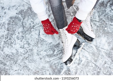 Girl tying shoelaces on ice skates before skating on the ice rink, hands in red knitted gloves. View from top.