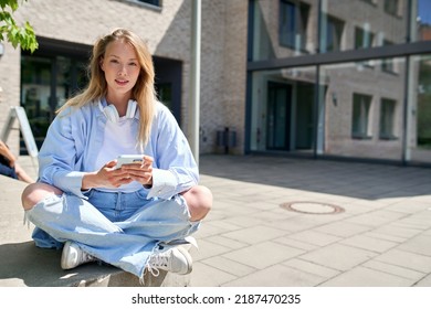 Girl university student wearing headphones holding cellphone using mobile apps on smartphone sitting on stairs outdoors looking at camera advertising online applications for education. Portrait - Powered by Shutterstock