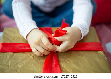 The Girl Unties A Red Bow On A Large Gold Gift Box.