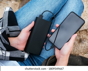 The girl uses a smartphone connected to the power Bank. Black smartphone and powerBank in women's hands close-up. Using gadgets in the modern world. The concept of portable chargers, modern tourism.