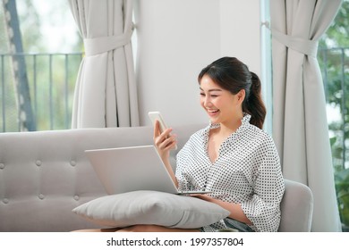 girl using laptop and smartphone. Young woman in casual clothes with her legs crossed sitting on sofa at cozy home interior. Technology and communication concept.