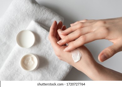 Girl using moisturizer. Smudge cream or body lotion on female hands in bathroom on white background with towel, indoors. Daily skincare routine vertical format, above