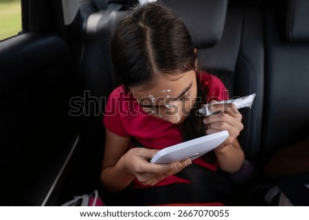 Girl using a small mirror while applying a medical cream on her face to treat chickenpox during a car trip