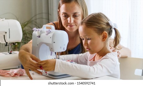 A girl is using a small sewing machine, a woman using a needle. A mother and a daughter are at home together, day time. Mommy explains a little child how to work with equipment and fabric. Close-up.