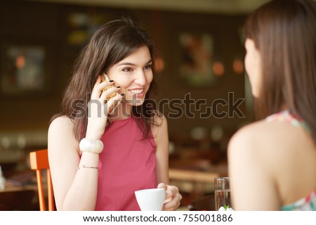 Girl using smart phone and drinking coffee while sitting with her friend in cafe