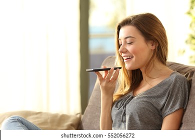 Girl using a smart phone voice recognition on line sitting on a sofa in the living room at home with a warm light and a window in the background