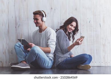 Girl using smartphone, and the guy using a tablet with headphones listening to music and leaning on each other, sit on wooden floor at home - Shutterstock ID 2102864569