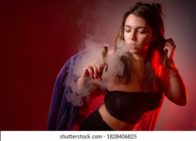 Girl With A VAPE On A Red Background. Vaping Concept. Vaper Girl In Black Clothing. A Woman In A Cloud Of Smoke From E-cigarette. 