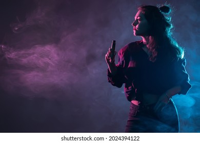 The girl with the vape. Smoking girl. Close-up of a woman inhaling an e-cigarette. The concept of night life. A woman with an e-cigarette in her hands on a dark smoky background. Space for text.