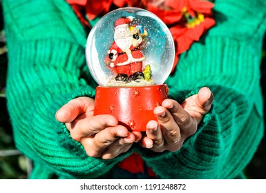 Girl in vibrant green knitted pullover and red Christmas decorations on her neck, holding big Christmas snow globe with happy Santa Claus inside and floating bright snow.