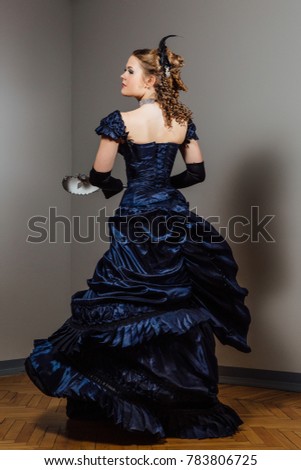 Girl in vintage blue dress with mask in hands. Actress in the role. Actor's game. Beautiful hairstyle. Decalte, open back.