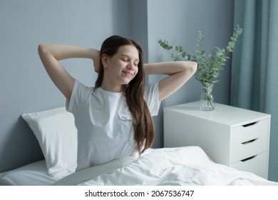 Girl waking up late in morning on bed and pillows, dressed in T-shirt, moving arms trying to get up, eyes closed, smiling