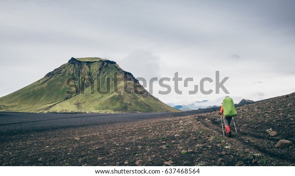 A girl is walking in the black obsidian\
desert of the third day of the Laugavegurinn hiking trail, Iceland,\
passing by one of the big green\
hills