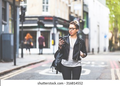 Girl walking down the street with her phone. A woman is walking alone in London. She is looking at her smart phone. Blurred on background there are  typical english houses and shops.