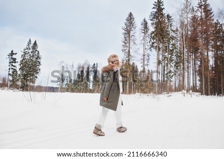 Girl walking in winter snow meadow, wearing stylish fashion outfit khaki parka coat jacket, fur hat and moon boots, full length, looking back. Happy smiling woman hiking in nature