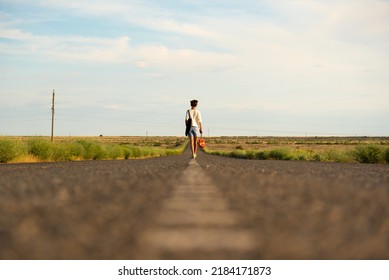 A Girl Walks Along An Empty Road In The Steppe. She Has A Ukulele In Her Hands And A Backpack On Her Shoulders. It's Summer And Warm Outside. Open Road.