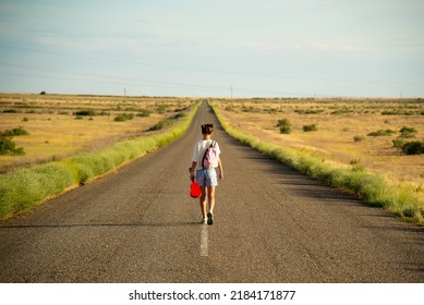 A Girl Walks Along An Empty Road In The Steppe. She Has A Ukulele In Her Hands And A Backpack On Her Shoulders. It's Summer And Warm Outside. Open Road.